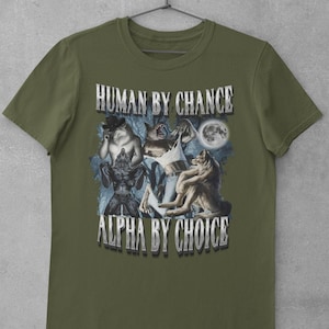 May include: An olive green short-sleeve t-shirt featuring a graphic print of three anthropomorphic wolves in a forest setting under a full moon. The text "HUMAN BY CHANCE" appears at the top and "ALPHA BY CHOICE" at the bottom in silver lettering.