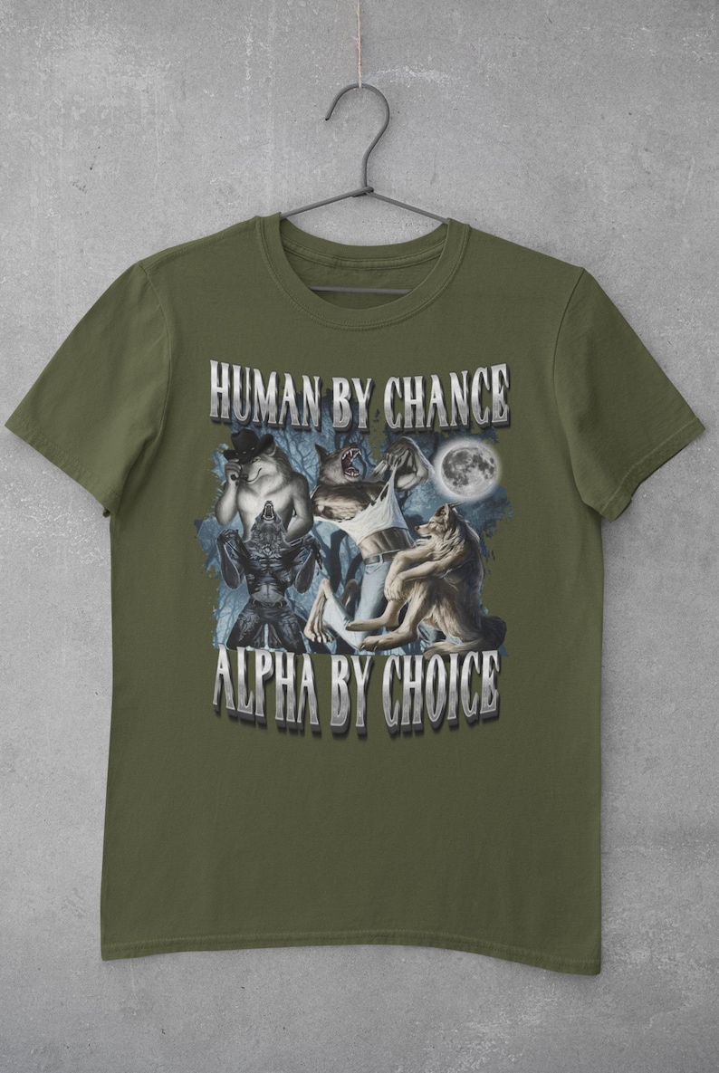 May include: An olive green short-sleeve t-shirt featuring a graphic print of three anthropomorphic wolves in a forest setting under a full moon. The text "HUMAN BY CHANCE" appears at the top and "ALPHA BY CHOICE" at the bottom in silver lettering.