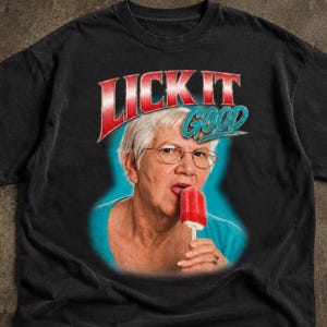 May include: A black t-shirt featuring a graphic print of an older person with short gray hair and glasses wearing a teal shirt. The person is eating a red popsicle. Above the image, the text LICK IT GOOD is printed in red and teal.