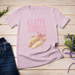 Puede incluir: Una camiseta rosa con el texto "Glizzy Gobbler" y un dibujo animado de un perrito caliente con mostaza y una cinta rosa.