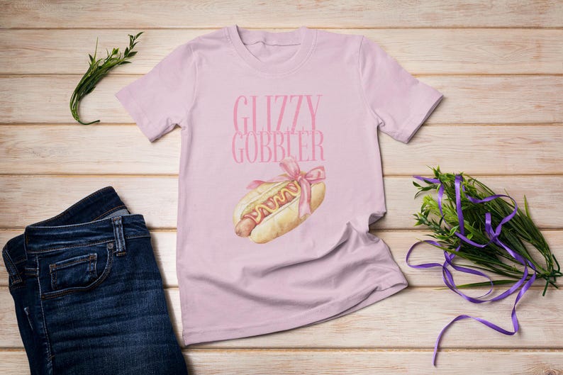 Puede incluir: Una camiseta rosa con el texto "Glizzy Gobbler" y un dibujo animado de un perrito caliente con mostaza y una cinta rosa.