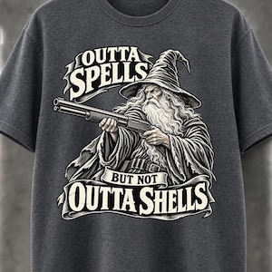 Puede incluir: Una camiseta gris carbón con una ilustración gráfica de un mago barbudo con sombrero puntiagudo que sostiene una escopeta. El texto OUTTA SPELLS BUT NOT OUTTA SHELLS aparece en una fuente estilizada sobre pancartas arriba y abajo.