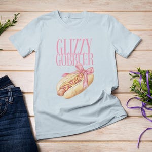 Puede incluir: Camiseta azul claro con el texto "Glizzy Gobbler" en letras rosas y un dibujo animado de un perrito caliente con mostaza y una cinta rosa en la parte superior.