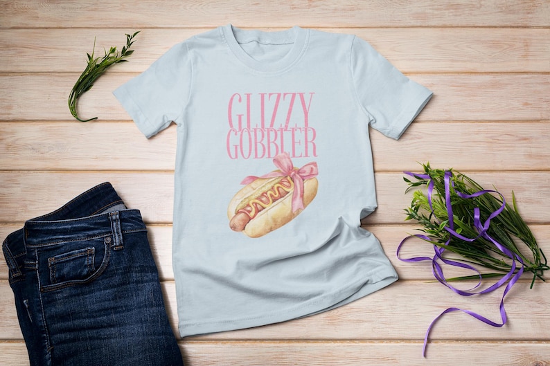 Puede incluir: Camiseta azul claro con el texto "Glizzy Gobbler" en letras rosas y un dibujo animado de un perrito caliente con mostaza y una cinta rosa en la parte superior.