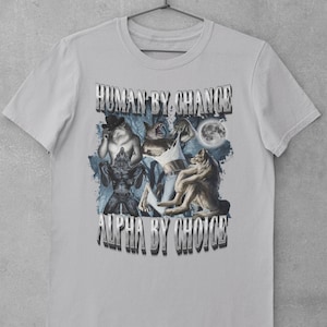 May include: A light gray short-sleeve t-shirt featuring a graphic print of multiple wolves and a full moon. The text "HUMAN BY CHANCE" appears at the top and "ALPHA BY CHOICE" at the bottom in a metallic, block-style font against a dark blue background.