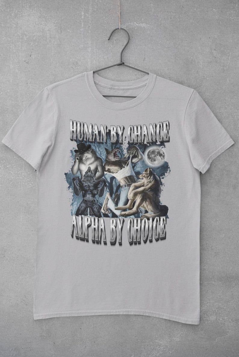 May include: A light gray short-sleeve t-shirt featuring a graphic print of multiple wolves and a full moon. The text "HUMAN BY CHANCE" appears at the top and "ALPHA BY CHOICE" at the bottom in a metallic, block-style font against a dark blue background.