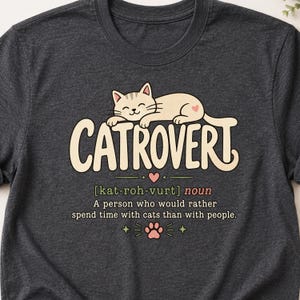 May include: A dark heather gray t-shirt featuring a graphic of a light tan cat resting above the text CATROVERT. Below is the phonetic spelling, the word noun, and the definition: A person who would rather spend time with cats than with people.