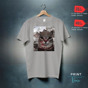 May include: A gray t-shirt with a print of a cat wearing a military helmet. The cat is looking directly at the viewer. The background of the print is a war scene with smoke and explosions. The text "35% OFF IF YOU BUY 3 ITEMS CODE: WAVE3" and "40% OFF IF YOU BUY 4 ITEMS CODE: WAVE4" is printed on the image. The text "PRINT Waave" is printed in blue on a white background at the bottom of the image.