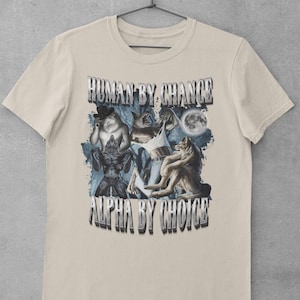 May include: A beige short-sleeve t-shirt featuring a graphic print of multiple wolves and a full moon. The text "HUMAN BY CHANCE" appears at the top and "ALPHA BY CHOICE" at the bottom in a metallic, block-style font. The design includes blue forest elements.
