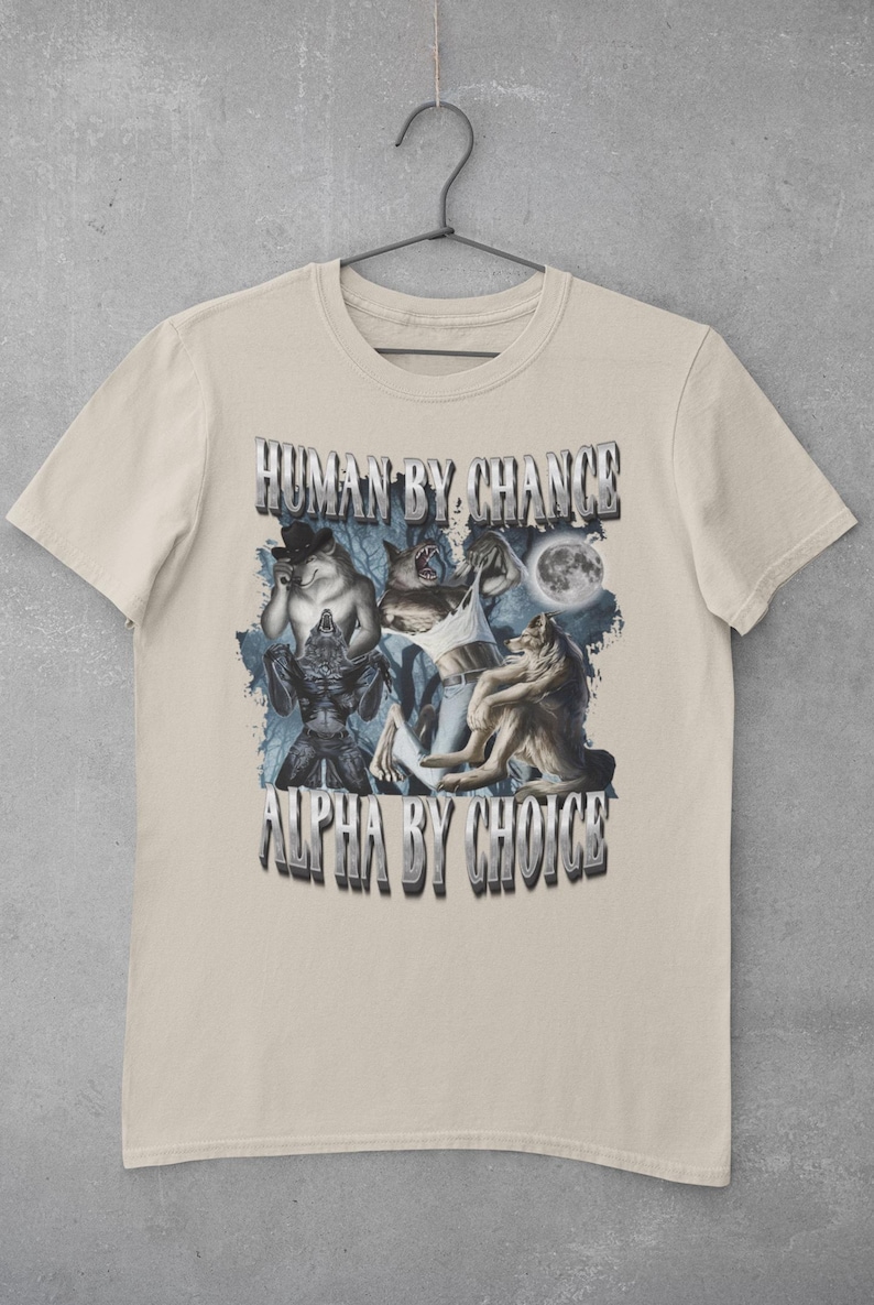 May include: A beige short-sleeve t-shirt featuring a graphic print of multiple wolves and a full moon. The text "HUMAN BY CHANCE" appears at the top and "ALPHA BY CHOICE" at the bottom in a metallic, block-style font. The design includes blue forest elements.