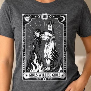 Può includere: Una t-shirt a maniche corte grigio scuro mélange con una stampa grafica bianca che raffigura una carta dei tarocchi. L'illustrazione mostra una persona legata a un palo con fiamme, il numero romano XIII e il testo GIRLS WILL BE GIRLS.