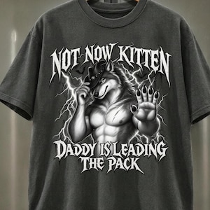 May include: A charcoal grey short-sleeve t-shirt featuring a graphic of a muscular wolf wearing a cowboy hat, surrounded by lightning bolts. The text reads NOT NOW KITTEN and DADDY IS LEADING THE PACK in a bold, stylised white gothic font.