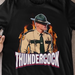 May include: Black t-shirt featuring a graphic of a man in a park ranger uniform with a green hat, set against a backdrop of flames. The word "THUNDERCOCK" is displayed in bold, white letters with red and blue accents.