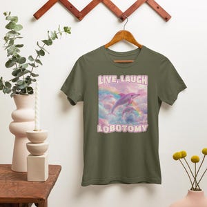 May include: A green t-shirt with a pink dolphin jumping over a rainbow in a cloud. The text "LIVE, LAUGH LOBOTOMY" is printed on the shirt.