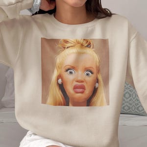 Könnte beinhalten: Ein cremefarbenes Sweatshirt mit Rundhalsausschnitt und einem quadratischen Grafikdruck auf der Brust. Der Druck zeigt ein Nahaufnahme-Porträt einer blonden Puppe mit überraschtem Gesichtsausdruck, großen Augen und vollen Lippen.