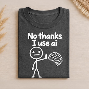 Pode incluir: Uma t-shirt cinzento-carvão escuro com um gráfico branco de uma figura de pau a levantar a mão para rejeitar uma ilustração de um cérebro. O texto acima diz "No thanks I use ai" numa fonte branca, arredondada e em negrito.