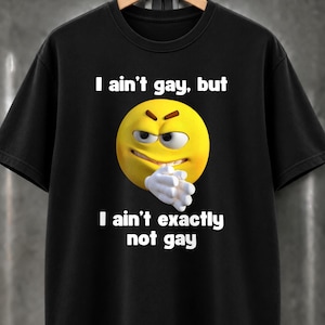 Könnte beinhalten: Ein schwarzes T-Shirt mit kurzen Ärmeln, das einen gelben Emoji mit schelmischem Gesichtsausdruck und gefalteten, weiß behandschuhten Händen zeigt. Der Text "I ain't gay, but I ain't exactly not gay" steht in weißen Buchstaben darüber und darunter.