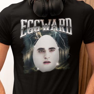 May include: Black t-shirt with the word "EGGWARD" in a silver, bold font. The shirt features a surreal image of a face inside an egg, set against a backdrop of lightning and a forest.
