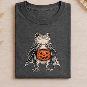 Op de afbeelding: Een donker antracietgrijs t-shirt met ronde hals en een grafische illustratie van een kikker verkleed als vampier, met een zwarte cape en hoektanden, die een oranje pompoen-emmer voor trick-or-treat vasthoudt.