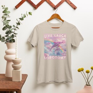 May include: A light beige t-shirt with a pink dolphin jumping over a rainbow. The text "LIVE, LAUGH LOBOTOMY" is printed on the shirt.