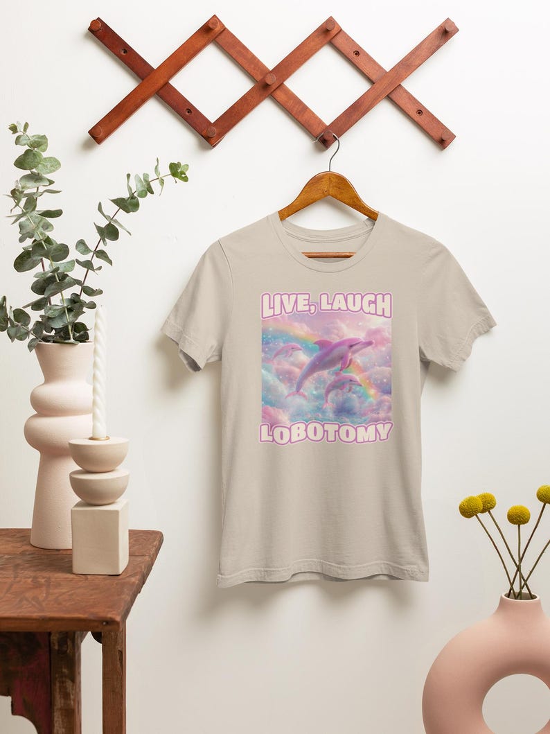 May include: A light beige t-shirt with a pink dolphin jumping over a rainbow. The text "LIVE, LAUGH LOBOTOMY" is printed on the shirt.