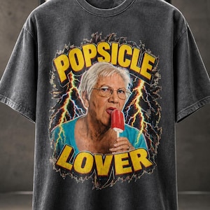 Può includere: Una t-shirt grigio antracite con una stampa grafica di una persona anziana con capelli bianchi corti e occhiali che mangia un ghiacciolo rosso. Il testo POPSICLE LOVER è stampato in grassetto giallo sopra e sotto l'immagine, circondato da fulmini gialli e blu su sfondo scuro.