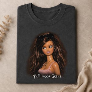 Könnte beinhalten: Ein anthrazitfarbenes T-Shirt mit einer Grafik einer Puppe mit langem braunem Haar, die eine schwarze Sonnenbrille auf dem Kopf und eine goldene Kreuzkette trägt. Die Puppe trägt ein rosa Oberteil. Der Text Y'all need Jesus steht darunter.
