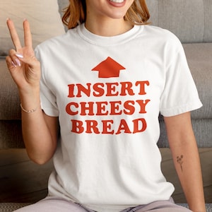 Puede incluir: Camiseta blanca con la frase "INSERT CHEESY BREAD" en rojo, con una flecha roja encima. La camiseta es de estilo casual, adecuada para el uso diario.