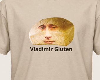 Può includere: Una t-shirt beige a maniche corte con una grafica centrale che raffigura una pagnotta con un volto umano. Sotto l'immagine del pane, il testo "Vladimir Gluten" è stampato in un carattere sans-serif di colore marrone scuro sul petto.