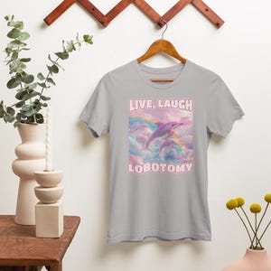 May include: A light gray t-shirt with a pink dolphin jumping over a rainbow with the text "LIVE, LAUGH, LOBOTOMY" in pink.