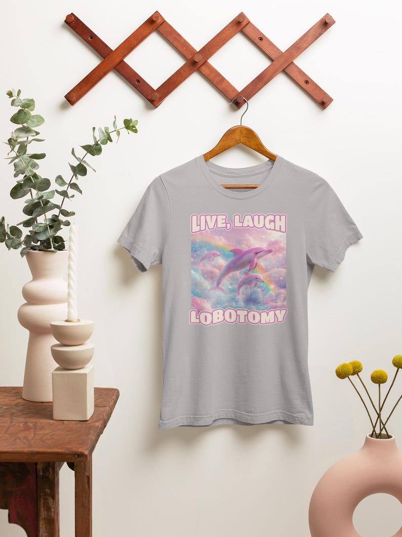 May include: A light gray t-shirt with a pink dolphin jumping over a rainbow with the text "LIVE, LAUGH, LOBOTOMY" in pink.