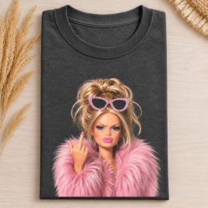 Puede incluir: Una camiseta gris carbón oscuro con un estampado gráfico de una muñeca con cabello rubio en un moño, usando gafas de sol de ojo de gato con pedrería rosa y un abrigo de piel sintética rosa, mientras hace un gesto con el dedo medio.