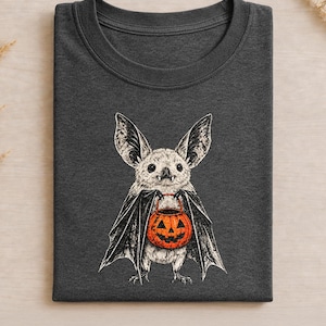 Puede incluir: Una camiseta de cuello redondo en color gris carbón oscuro con una ilustración gráfica detallada de un murciélago claro con orejas grandes y colmillos. El murciélago sostiene un cubo de calabaza naranja brillante con una cara tallada.