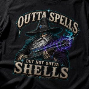 Könnte beinhalten: Ein schwarzes T-Shirt mit einer Grafik eines Zauberers mit langem weißem Bart und spitzem Hut, der eine doppelläufige Schrotflinte hält, die violette magische Energie ausstrahlt. Der Text lautet OUTTA SPELLS BUT NOT OUTTA SHELLS in fetter Schrift.