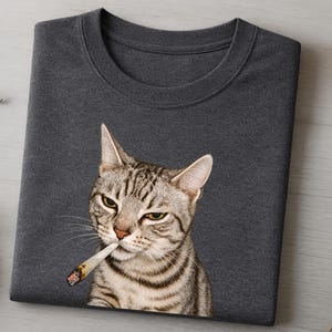 May include: A dark heather grey crew neck t-shirt featuring a graphic print of a tabby cat with half-closed eyes holding a lit cigarette in its mouth. The cat has grey and black stripes, pointed ears, and long white whiskers against the dark fabric.