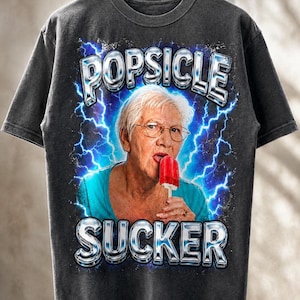 Könnte beinhalten: Ein anthrazitfarbenes Kurzarm-T-Shirt mit einem Grafikdruck einer älteren Person mit Brille, die ein rotes Eis am Stiel isst. Das Design zeigt blaue Blitze im Hintergrund und den metallisch wirkenden Schriftzug POPSICLE SUCKER.