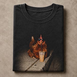 Op de afbeelding: Een zwart t-shirt met ronde hals en een grafische print van een bruine kip die een zwarte zonnebril draagt en drinkt uit een doorzichtige plastic beker met een rietje, zittend op een grijze betonnen rand tegen een donkere achtergrond.