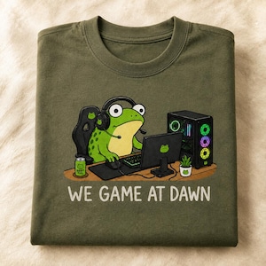 Könnte beinhalten: Ein olivgrünes T-Shirt mit einer Grafik eines grünen Frosches, der schwarze Kopfhörer trägt und in einem Gaming-Stuhl an einem Schreibtisch mit Computer, Tastatur, Maus, einer Dose mit der Aufschrift Ribbit Fuel und einer kleinen Pflanze sitzt, mit dem Text WE GAME AT DAWN.
