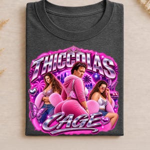 May include: A dark gray t-shirt featuring a graphic print with the text "THICCOLAS CAGE". The design includes a central figure in a pink velour tracksuit flanked by two individuals, surrounded by pink butterflies, hearts, and palm trees.