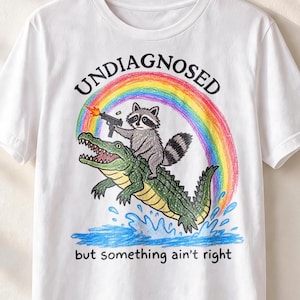 Könnte beinhalten: Ein weißes T-Shirt mit einer bunten Buntstiftzeichnung eines Waschbären, der auf einem grünen Alligator reitet. Der Waschbär hält eine feuernde Waffe. Darüber ist ein Regenbogen und der Text UNDIAGNOSED, darunter der Text but something ain't right.