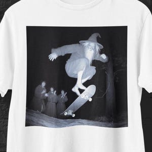Könnte beinhalten: Ein weißes T-Shirt mit einem schwarz-weißen Grafikdruck eines Zauberers mit langem Bart und spitzem Hut, der auf einem Skateboard springt, während drei in Roben gekleidete Gestalten im Hintergrund zuschauen.