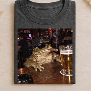 Könnte beinhalten: Ein dunkelgraues T-Shirt mit Rundhalsausschnitt und einem Grafikdruck einer Kröte, die eine schwarze Baseballkappe und eine dunkle Sonnenbrille trägt, eine Zigarre raucht und an einer Holztheke neben einem Bierglas und Aschenbecher sitzt.