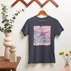 May include: A navy blue t-shirt with a pink graphic of two dolphins jumping over a rainbow in the clouds. The text "LIVE, LAUGH, LOBOTOMY" is printed in pink on the shirt.
