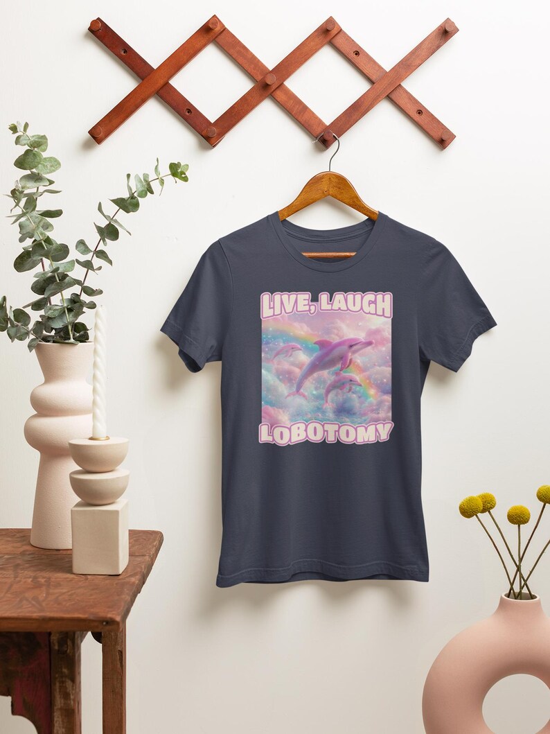 May include: A navy blue t-shirt with a pink graphic of two dolphins jumping over a rainbow in the clouds. The text "LIVE, LAUGH, LOBOTOMY" is printed in pink on the shirt.