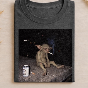 May include: A dark charcoal gray crew neck t-shirt featuring a graphic print of a small, wrinkled, green creature with large ears sitting on a ledge, smoking a cigarette, with a silver beverage can and a discarded cigarette butt nearby.