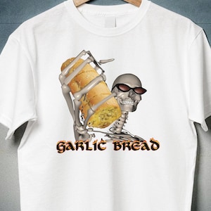 May include: A white short-sleeve t-shirt featuring a graphic of a skeleton wearing black sunglasses and holding a loaf of garlic bread. Below the skeleton, the text GARLIC BREAD is printed in a stylized font with a fiery orange and yellow border.