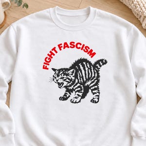 Op de afbeelding: Een wit sweatshirt met lange mouwen en een zwarte afbeelding van een blazend, getijgerd kitten met een gebogen rug. Boven het kitten staat de tekst FIGHT FASCISM in dikke, rode hoofdletters. Het kledingstuk heeft een ronde hals en boorden.