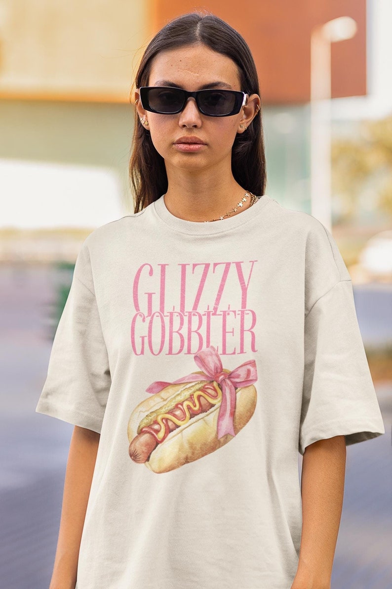 Puede incluir: Una camiseta blanca con un gr&aacute;fico rosa de un perrito caliente con un lazo rosa y el texto "GLIZZY GOBBLER".