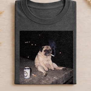 Könnte beinhalten: Ein dunkelgraues T-Shirt mit Rundhalsausschnitt und einem Grafikdruck eines beigen Mopses, der nachts auf einem Betonvorsprung sitzt. Der Mops hat eine brennende Zigarette im Maul mit sichtbarem Rauch und eine silberne Aluminiumdose steht daneben.