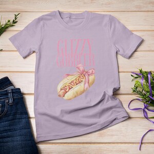 Puede incluir: Una camiseta lavanda con el texto "Gizzy Gourmet" en letras rosas. Una caricatura de un hot dog con mostaza y un lazo rosa est&aacute; impresa en la camiseta.